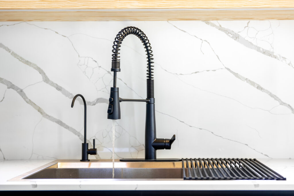 Water flows from a black professional faucet over a steel sink against a white marble wall.